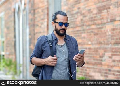 people, technology, travel and tourism - man with earphones, smartphone and bag walking along city street and listening to music