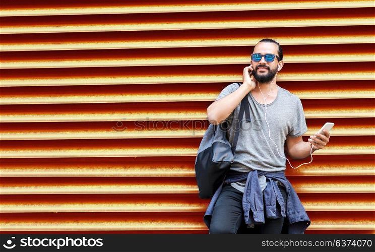 people, technology, travel and tourism - man with earphones, smartphone and bag on city street and listening to music over ribbed red wall background. man with earphones and smartphone over wall