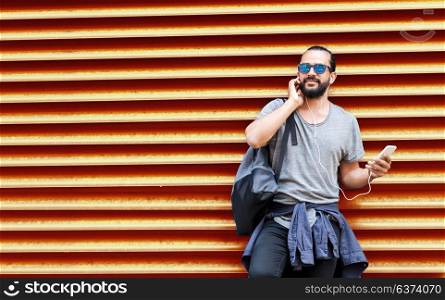 people, technology, travel and tourism - man with earphones, smartphone and bag on city street and listening to music over ribbed red wall background. man with earphones and smartphone over wall