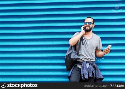 people, technology, travel and tourism - man with earphones, smartphone and bag on city street and listening to music over ribbed blue wall background. man with earphones and smartphone over wall