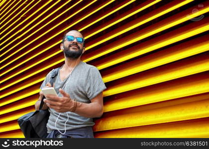 people, technology, travel and tourism - man with earphones, smartphone and bag on city street and listening to music over ribbed yellow wall background. man with earphones and smartphone over wall