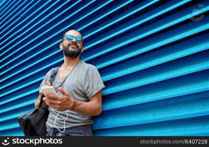 people, technology, travel and tourism - man with earphones, smartphone and bag on city street and listening to music over ribbed blue wall background. man with earphones and smartphone over wall