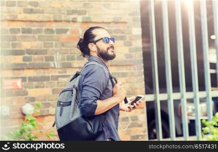 people, technology, travel and tourism - man with earphones, smartphone and bag walking along city street and listening to music . man with earphones and smartphone walking in city. man with earphones and smartphone walking in city