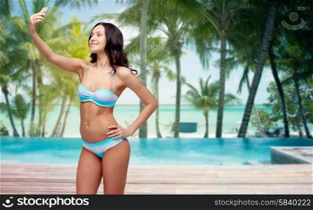 people, technology, travel, tourism and summer concept - happy young woman in bikini swimsuit taking selfie with smatphone over swimming pool and beach with palm trees background