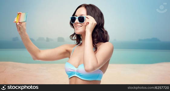 people, technology, travel, tourism and summer concept - happy young woman in bikini swimsuit and sunglasses taking selfie with smatphone over infinity pool at sea side background