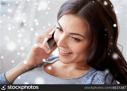 people, technology, winter, christmas and communication concept - happy young woman calling on smartphone at home over snow