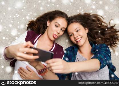 people, technology, winter, christmas and friendship concept - happy smiling pretty teenage girls or friends lying on floor and taking selfie with smartphone over snow