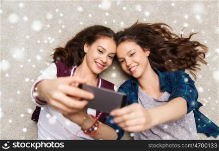 people, technology, winter, christmas and friendship concept - happy smiling pretty teenage girls or friends lying on floor and taking selfie with smartphone over snow