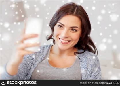 people, technology, winter, christmas and leisure concept - happy woman taking selfie with smartphone at home over snow