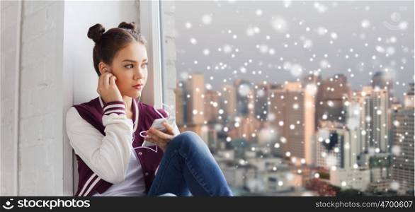 people, technology, winter, christmas and teens concept - sad pretty teenage girl sitting at window with smartphone and earphones listening to music over city background over snow