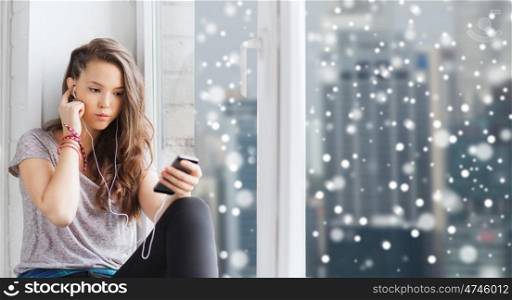 people, technology, winter, christmas and teens concept - sad pretty teenage girl sitting at window with smartphone and earphones listening to music over city background over snow