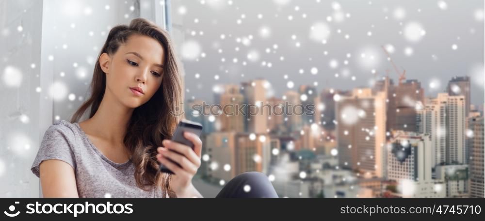people, technology, winter, christmas and teens concept - sad unhappy pretty teenage girl sitting at window with smartphone and texting over snow