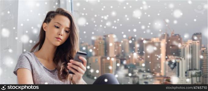 people, technology, winter, christmas and teens concept - sad unhappy pretty teenage girl sitting at window with smartphone and texting over snow