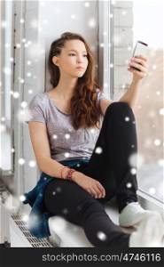 people, technology, winter, christmas and teens concept - sad unhappy pretty teenage girl sitting on windowsill with smartphone and texting over snow