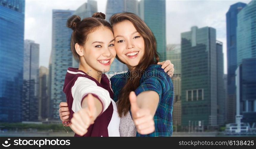 people, teens, friendship, travel and tourism concept concept - happy smiling pretty teenage girls hugging and showing thumbs up over singapore city background