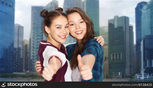 people, teens, friendship, travel and tourism concept concept - happy smiling pretty teenage girls hugging and showing thumbs up over singapore city background