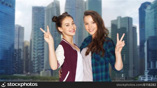 people, teens, friendship, travel and tourism concept - happy smiling pretty teenage girls hugging and showing peace hand sign over singapore city background