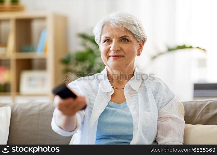 people, television and entertainment concept - senior woman in eyeglasses with remote control watching tv at home. senior woman with remote watching tv at home