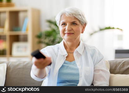 people, television and entertainment concept - senior woman in eyeglasses with remote control watching tv at home. senior woman with remote watching tv at home