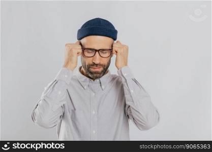 People, tension and migraine concept. Depressed unshaven young male suffers from headache, keeps hands in fists near temples, closes eyes, tries to concentrate, stands against white background