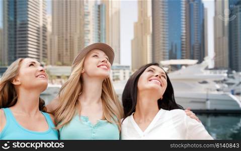 people, tourism, travel and vacation concept - happy young women looking up over dubai city harbour or waterfront with boats background