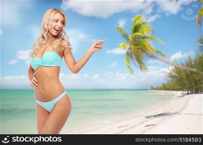 people, travel and summer holidays concept - happy smiling young woman in bikini swimsuit pointing finger over exotic tropical beach with palm trees background. happy smiling young woman in bikini on beach