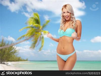 people, travel and summer holidays concept - happy smiling young woman in bikini swimsuit doing fist pump gesture over exotic tropical beach with palm trees background. happy woman in bikini doing fist pump on beach