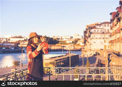 People, travel, holidays and adventure concept. Young woman with long hair walking on city street at sunrise, wearing hat and coat, making selfie with smartphone. Young woman with long hair walking on city street at sunrise, wearing hat and coat, making selfie with smartphone