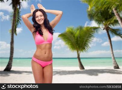 people, travel, summer and sexual concept - happy young woman in pink bikini swimsuit making bunny ears gesture over tropical beach with palm trees background