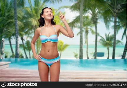 people, travel, swimwear and summer concept - happy young woman in bikini swimsuit pointing finger up to something imaginary over tropical beach with palm trees and pool at hotel resort background
