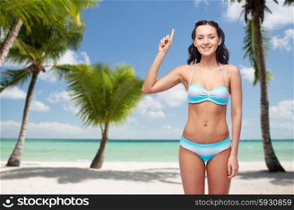 people, travel, swimwear and summer concept - happy young woman in bikini swimsuit pointing finger up to something imaginary over tropical beach with palm trees background