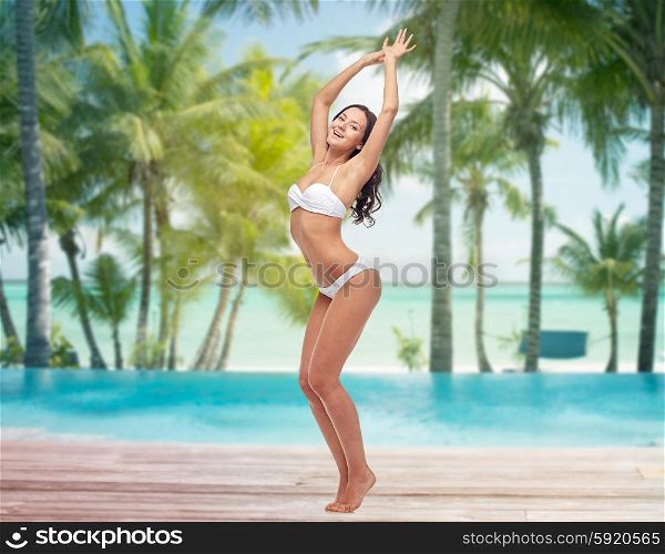 people, travel, swimwear and summer concept - happy young woman posing in white bikini swimsuit dancing with raised hands over tropical beach with palm trees and pool at hotel resort background