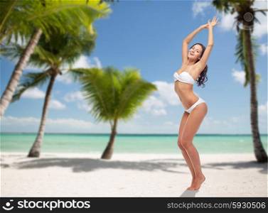 people, travel, swimwear and summer concept - happy young woman posing in white bikini swimsuit dancing with raised hands over tropical beach with palm trees background