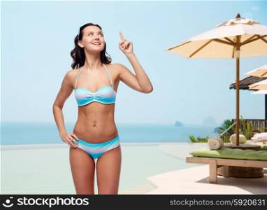 people, travel, swimwear, summer and beach concept - happy young woman in bikini swimsuit pointing finger and looking up to something imaginary over infinity edge pool at hotel resort background