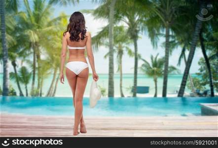 people, travel, swimwear, summer and beauty concept - young woman in white bikini swimsuit from back over tropical beach with palm trees and pool at hotel resort background