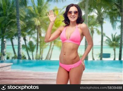 people, travel, swimwear, summer and gesture concept - happy young woman in sunglasses and pink swimsuit waving hand over tropical beach with palm trees and pool at hotel resort background