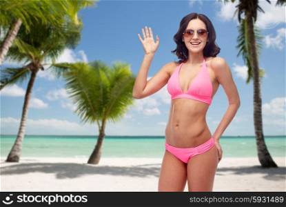 people, travel, swimwear, summer and gesture concept - happy young woman in sunglasses and pink swimsuit waving hand over tropical beach with palm trees background