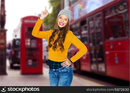 people, travel, tourism and fashion concept - happy young woman or teen girl in casual clothes and hipster hat pointing finger up over london city street background