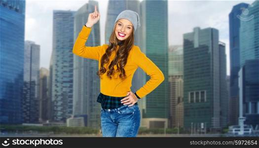 people, travel, tourism and fashion concept - happy young woman or teen girl in casual clothes and hipster hat pointing finger up over city buildings background