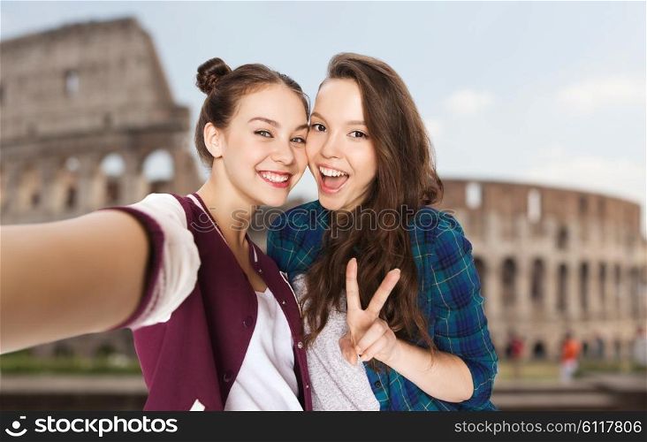 people, travel, tourism and friendship concept - happy smiling pretty teenage girls taking selfie and showing peace sign over coliseum in rome background