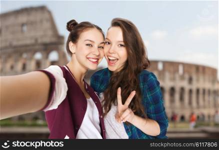 people, travel, tourism and friendship concept - happy smiling pretty teenage girls taking selfie and showing peace sign over coliseum in rome background
