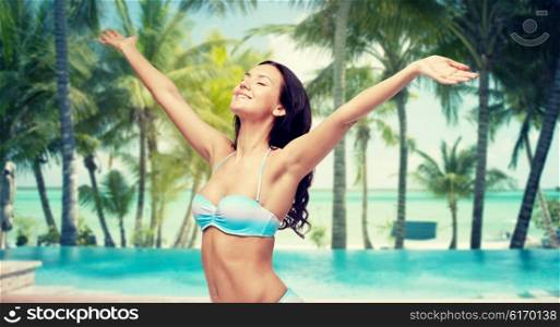 people, travel, tourism and summer concept - happy young woman in bikini swimsuit with raised hands looking up over swimming pool and beach with palm trees background
