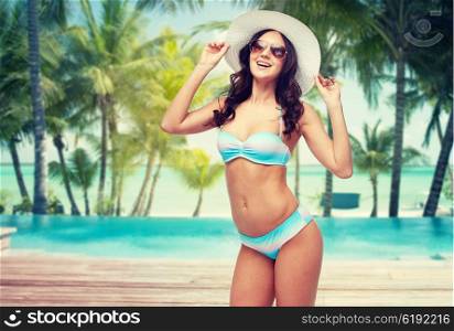 people, travel, tourism and summer concept - happy young woman in bikini swimsuit, sunglasses and sun hat over swimming pool and beach with palm trees background