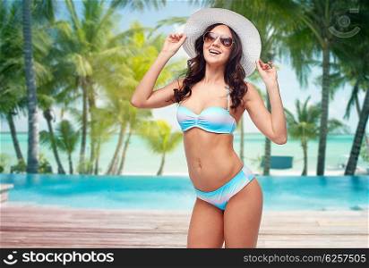 people, travel, tourism and summer concept - happy young woman in bikini swimsuit, sunglasses and sun hat over swimming pool and beach with palm trees background