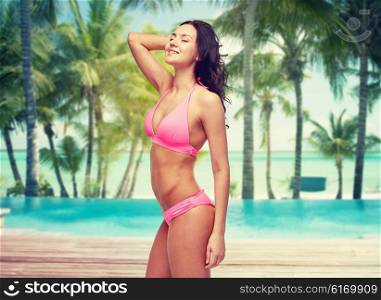 people, travel, tourism and summer concept - happy young woman posing in pink bikini swimsuit over swimming pool and beach with palm trees background