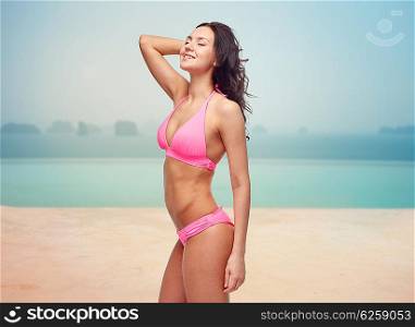 people, travel, tourism and summer concept - happy young woman posing in pink bikini swimsuit over infinity pool at sea side background