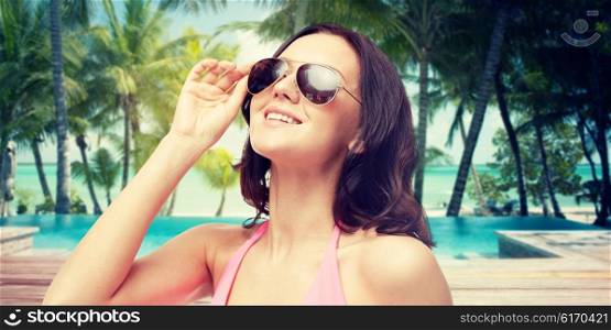 people, travel, tourism and summer resort concept - happy young woman in sunglasses looking up over swimming pool and beach with palm trees background
