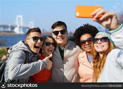 people, travel, tourism, friendship and technology concept - group of happy teenage friends taking selfie with smartphone and showing thumbs up over rainbow bridge at tokyo in japan background