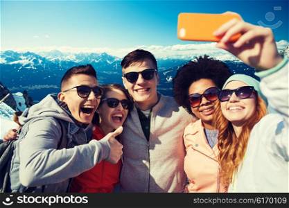 people, travel, tourism, friendship and technology concept - group of happy teenage friends taking selfie with smartphone and showing thumbs up over alps mountains in austria background