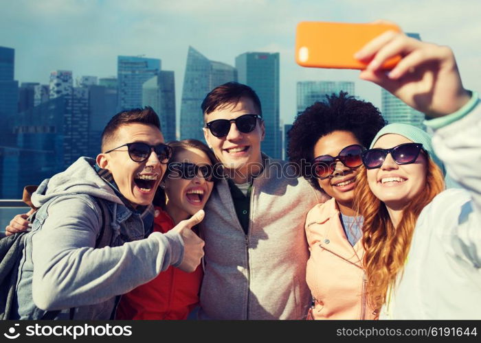 people, travel, tourism, friendship and technology concept - group of happy teenage friends taking selfie with smartphone and showing thumbs up over singapore city background
